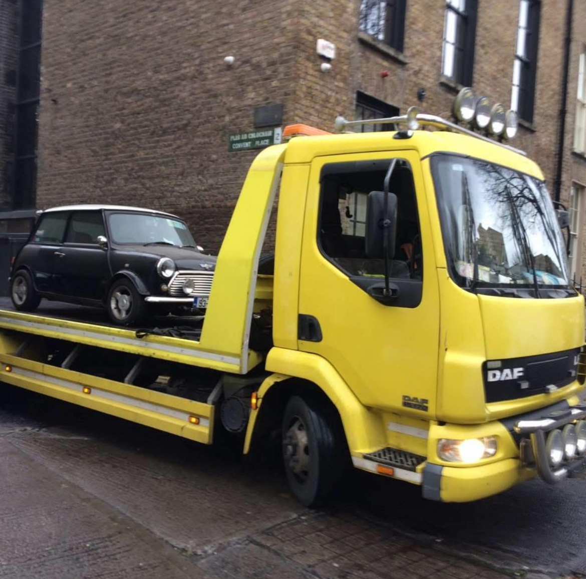 Yellow DAF flatbed tow truck from Tow&Go carrying a classic Mini Cooper on a city street
